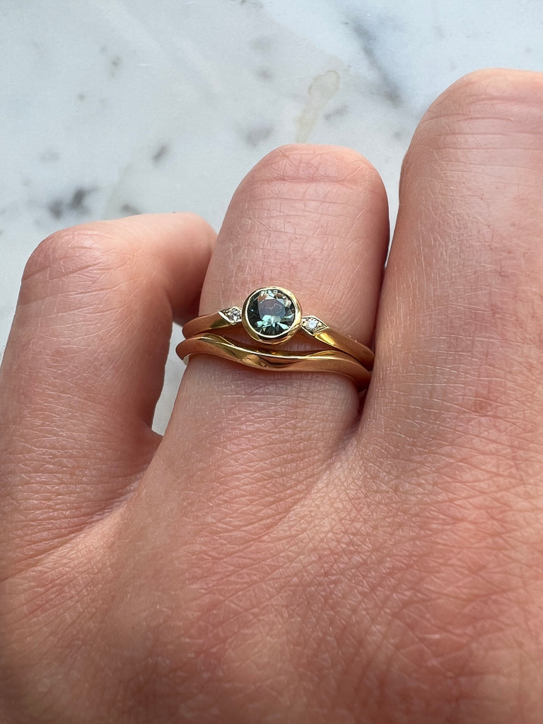 Round teal coloured sapphire and diamond engagement ring being worn with a curved wedding ring 